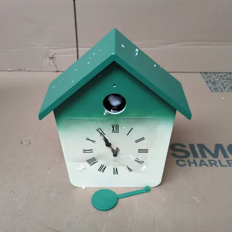 MODERN CUCKOO CLOCK FEATURES A WHIMSICAL HOUSE-SHAPED DESIGN WITH A VIBRANT GREEN ROOF AND A MATCHING PENDULUM