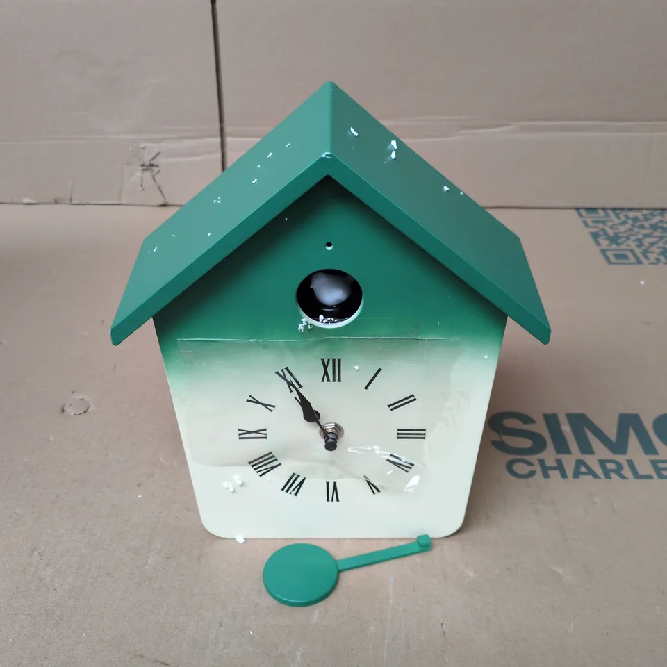 MODERN CUCKOO CLOCK FEATURES A WHIMSICAL HOUSE-SHAPED DESIGN WITH A VIBRANT GREEN ROOF AND A MATCHING PENDULUM